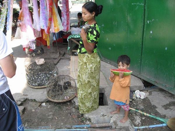 A girl sells sparrows & a bewildered-looking owl, while her little sister enjoys some water-melon.