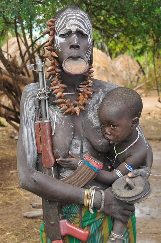 Mursi woman with lip plate, suckling babe and automatic weapon - Mago National Park, Omo Valley, Ethiopia