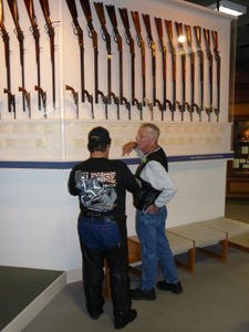 A man wearing chaps discusses guns with his friend
