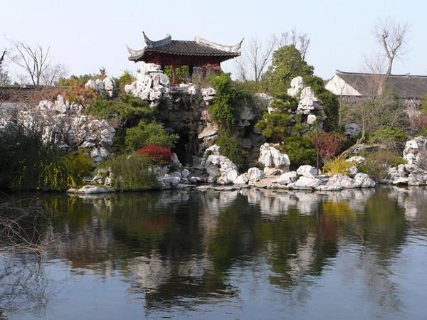 Een vijver, een berglandschap en een pagode horen er altijd bij in een klassieke Chinese tuin. Evenals een brug waarvan je nog net een stukje leuning ziet.
