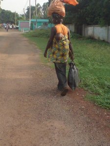 Lady carrying a bag on her head and  a baby on her back.
