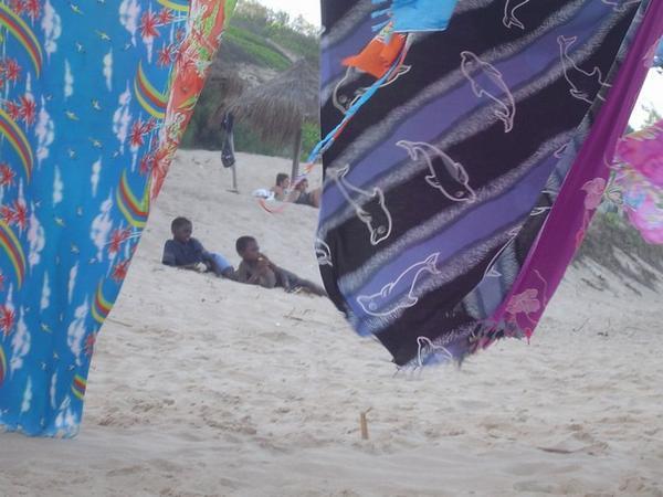 Beautiful sarrongs hang from a string in the beach. At the back you can see the kids just chilling out..  a common scene