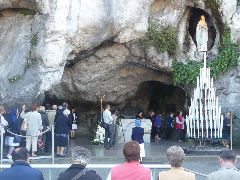 Pilgrims filing through the Grotte de Massabielle in silence, touching the walls with their left hands as they progress