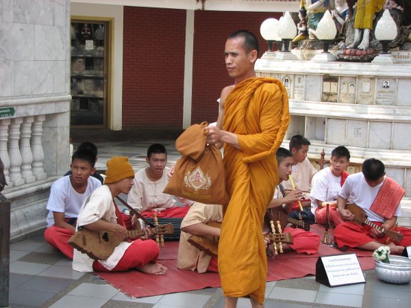 Tourist  monk walking by a group of teenage boys playing Thai music with various kinds of Thai music instruments.