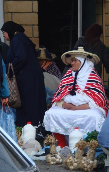 Berber woman selling produce at the markets in Tangier while her husband drinks tea with his friends
