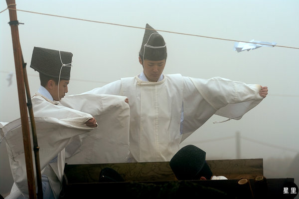Two Shinto priests are using a bow drill to light a fire while two others are trying to keep them out of the strong winds.